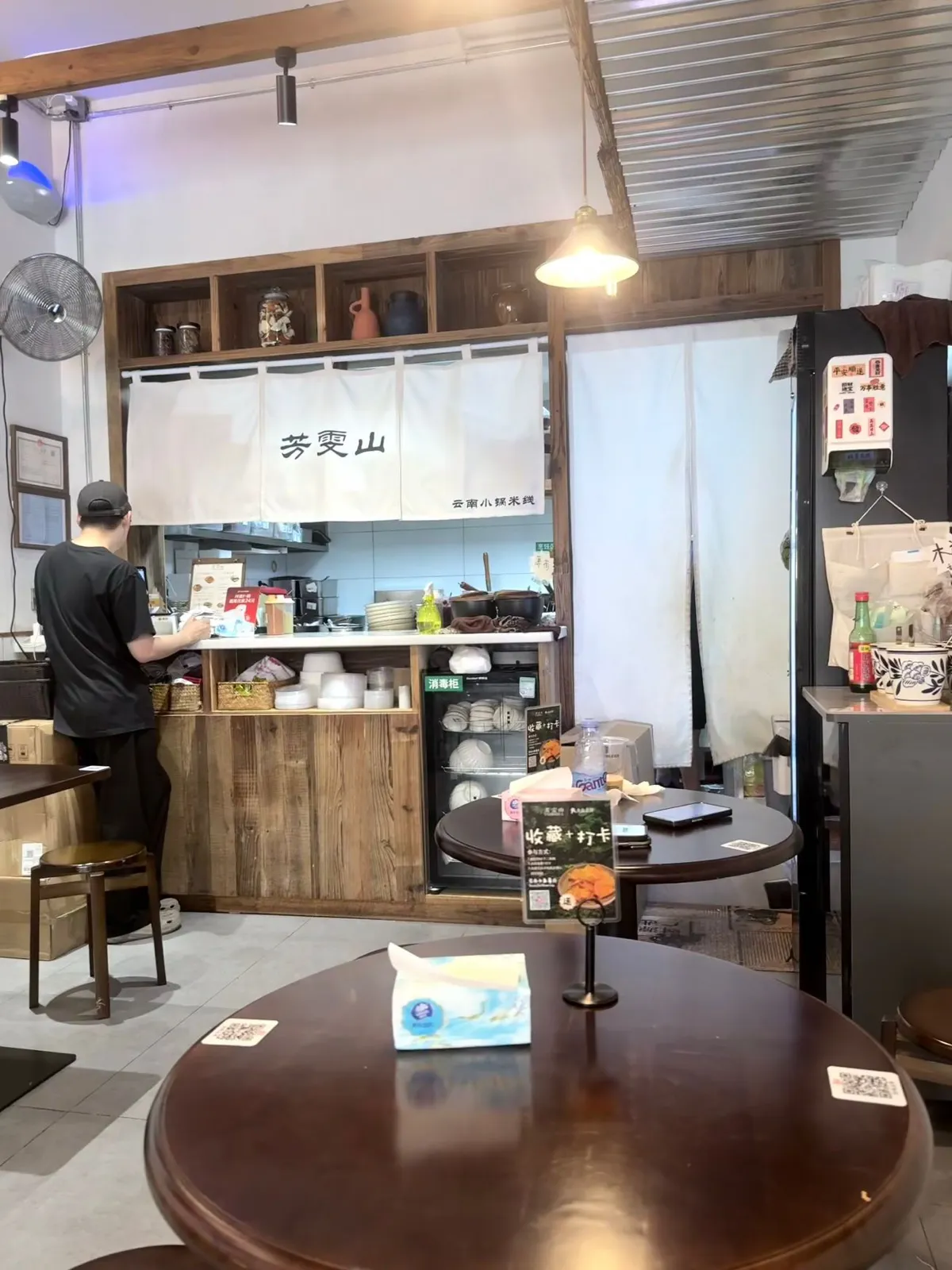 店內望向開放式廚房：木框吧枱後面掛住白色暖簾，寫住「芳雯山」，右下角細字「云南小锅米线」；枱面幾隻煮鍋、疊起嘅碗、消毒櫃，一位戴鴨舌帽嘅人背向鏡頭喺收銀位埋單；前景係圓木枱同紙巾