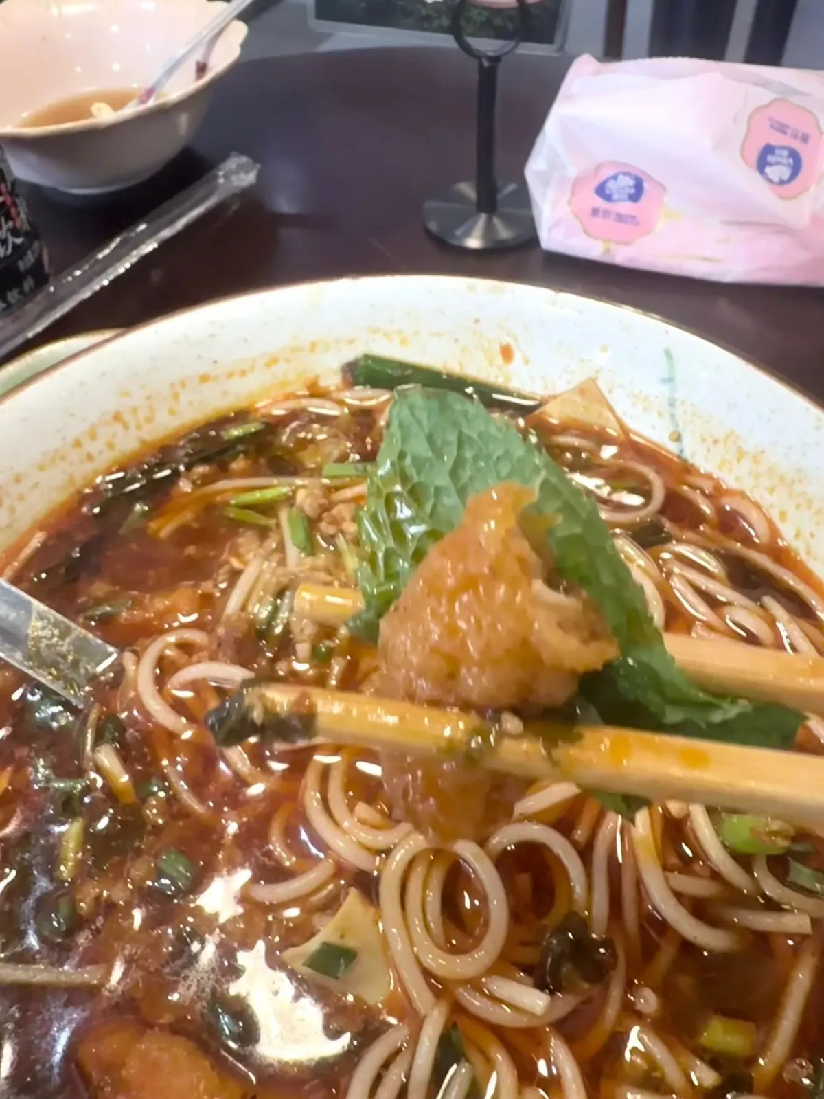小鍋米線特寫：紅油辣湯入面浮住兩塊完整嘅薄荷葉，葉面有明顯皺褶同鋸齒邊，同旁邊細碎嘅蔥花香菜好唔同；筷子夾住一件煎到金黃嘅酥肉，下面係白色米線、肉碎同豆皮