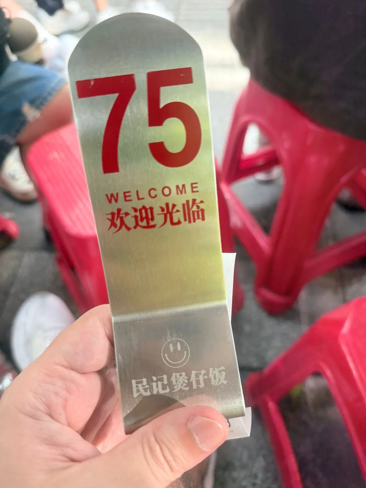 一隻手揸住民記煲仔飯嘅金屬派籌牌，紅色大字寫住 75，下面印住 WELCOME、欢迎光临同店名；背景係行人路上排開嘅紅色膠凳，同幾對等緊位嘅腳