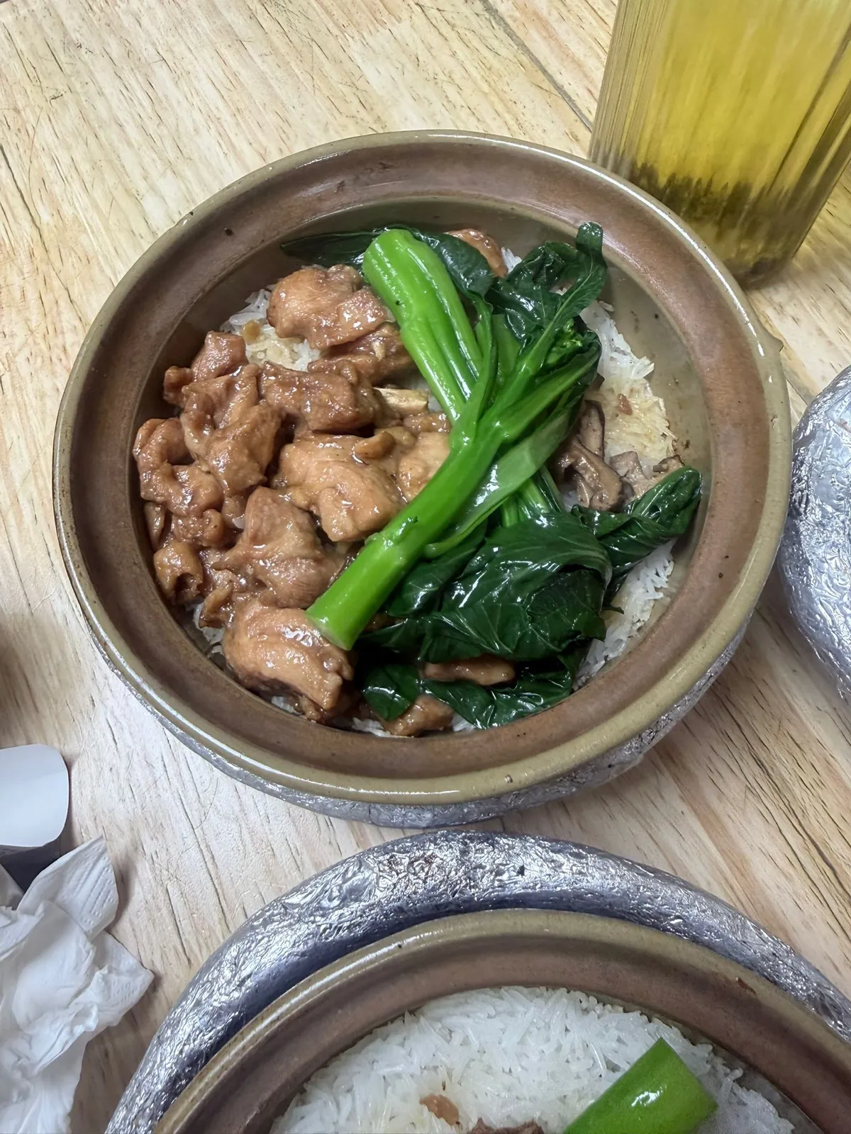 打開咗蓋嘅冬菇滑雞煲仔飯，雞件掛住淺啡色芡汁，旁邊有兩三片冬菇同一棵菜心，露出嘅米飯同樣係白色；右上角有杯黃色飲品，鏡頭下方係另一煲