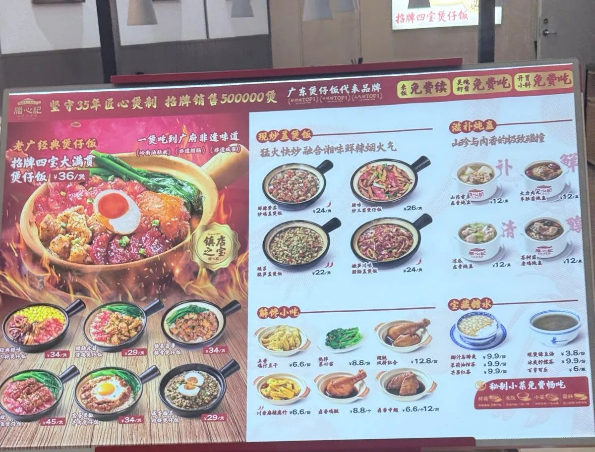 立喺店外嘅紅白色價目立牌：左半版係招牌四寶大滿貫煲仔飯嘅大圖，旁邊標住每煲價錢，下面排住六款煲仔飯嘅細圖同價；右半版分四格列出現炒蓋煲飯、燉盅、小吃同糖水，每項有相同價錢