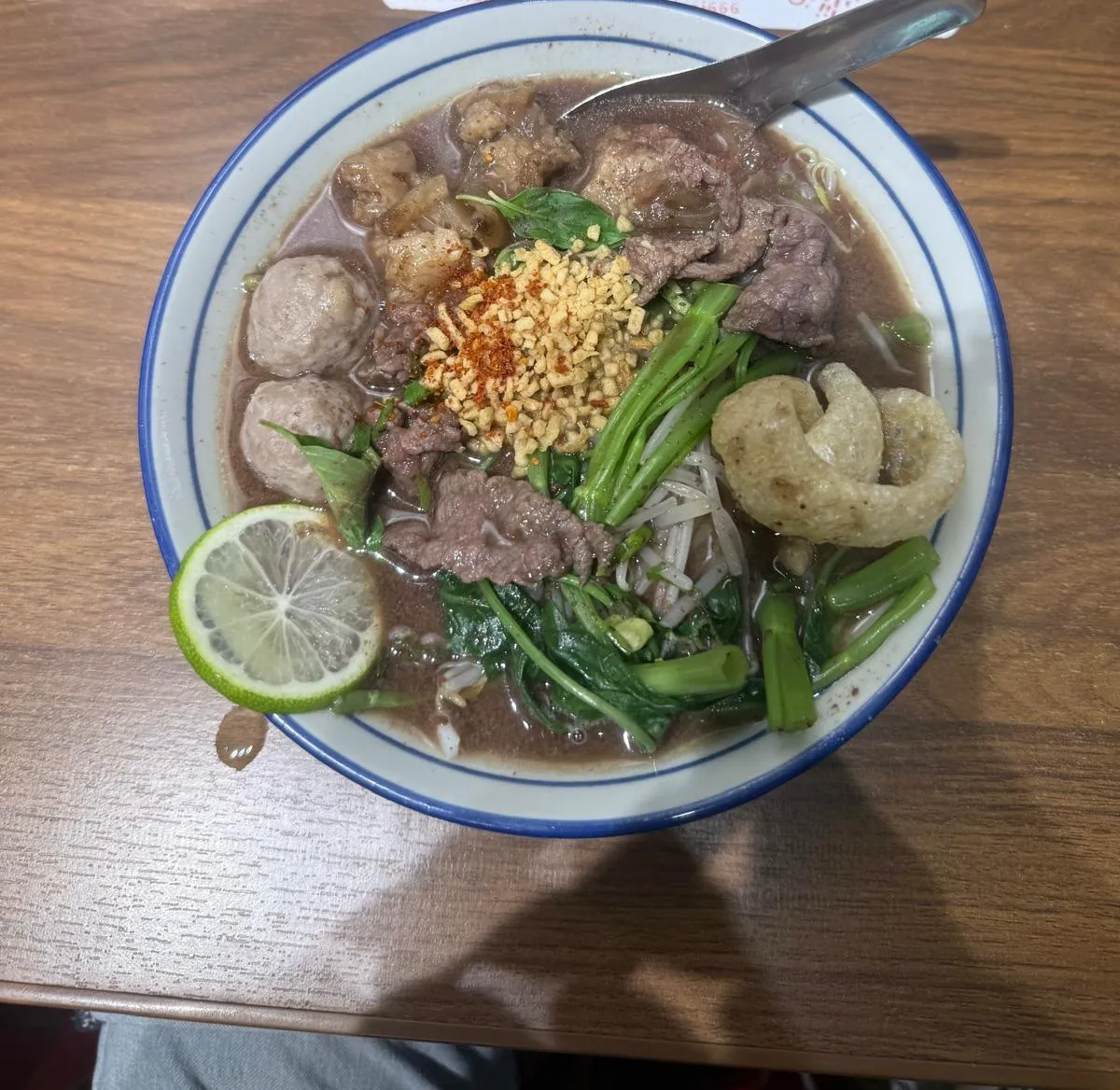 藍邊白瓷碗盛住嘅泰式牛肉船面：深啡色濃湯浸住薄切牛肉片、兩粒牛肉丸同軟腍牛腩，中央堆咗一撮炸蒜蓉同辣椒粉，旁邊有芥蘭、豆芽、九層塔同一塊炸豬皮，碗邊放咗一片青檸