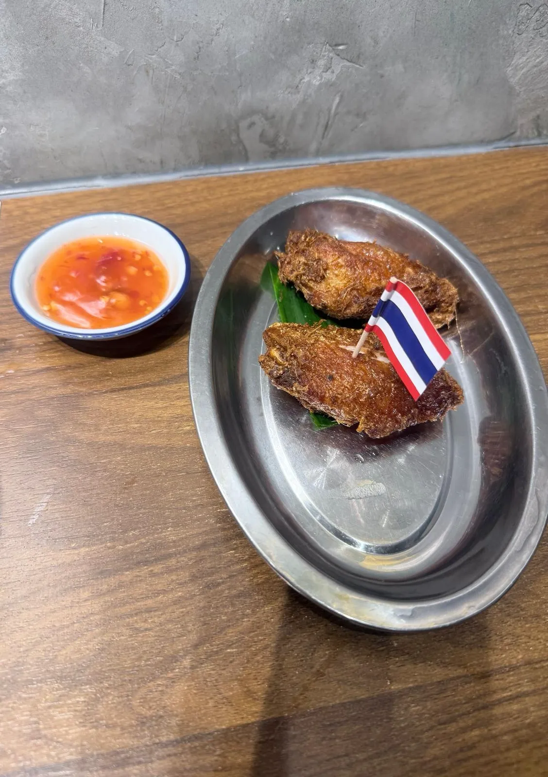 不鏽鋼橢圓碟上兩隻炸雞翼，外皮金黃起皺，插住一支細泰國國旗牙籤，底下墊住班蘭葉；旁邊一隻藍邊白瓷小碟盛住橙紅色泰式甜辣醬，入面見到蒜蓉同辣椒碎