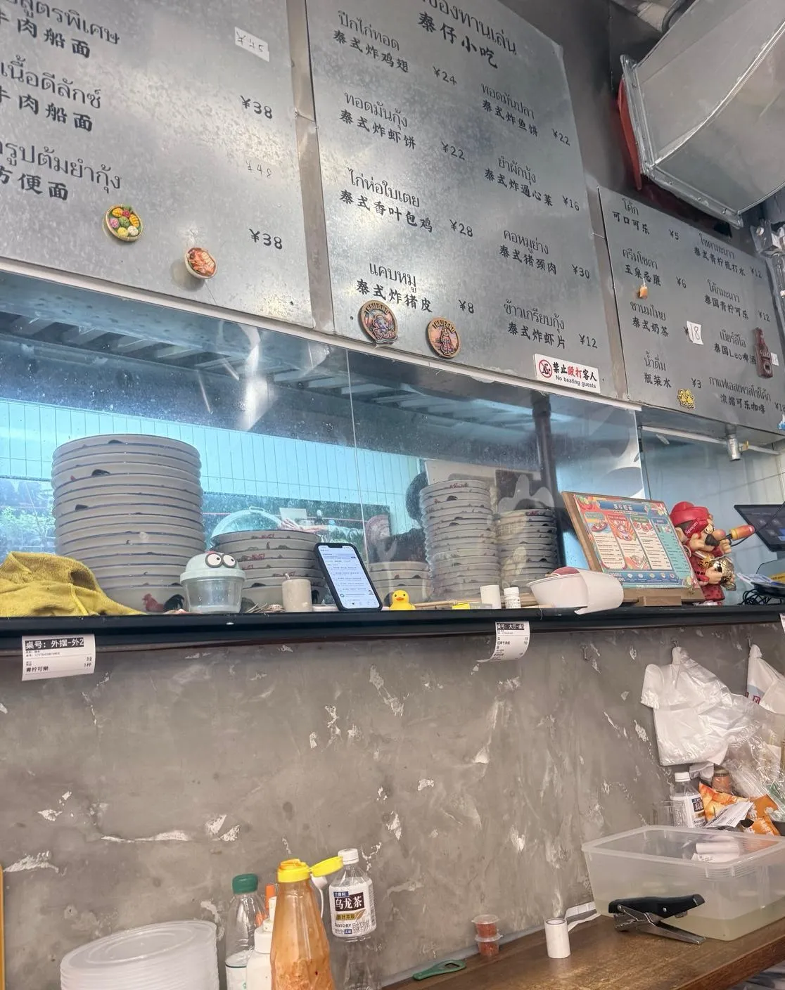 開放式廚房嘅出餐口：上方掛住幾塊金屬價目牌，泰文同中文雙語逐行列出菜名同價錢；玻璃後面疊住幾十隻船面碗，枱面有平板點餐機同醬料樽，下方係一條放住膠杯、辣椒醬同外賣袋嘅工作枱