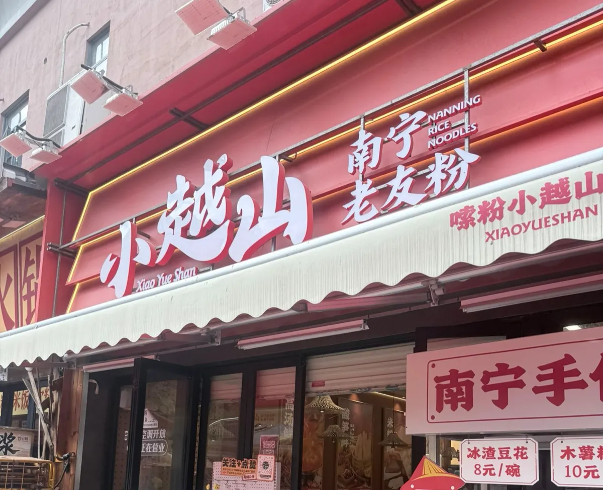 街舖招牌：紅底白色大字「小越山 Xiao Yue Shan」，右邊紅字「南寧老友粉」同英文 NANNING RICE NOODLES，再右邊淺色布幕上印住「嗦粉小越山 XIAOYUESHAN」；招牌下面係波浪邊遮篷同玻璃趟門，門邊貼住「空調開放 正在營業」，右邊掛住一塊白底紅字價目牌，寫住冰渣豆花 8 元／碗、木薯粉 10 元