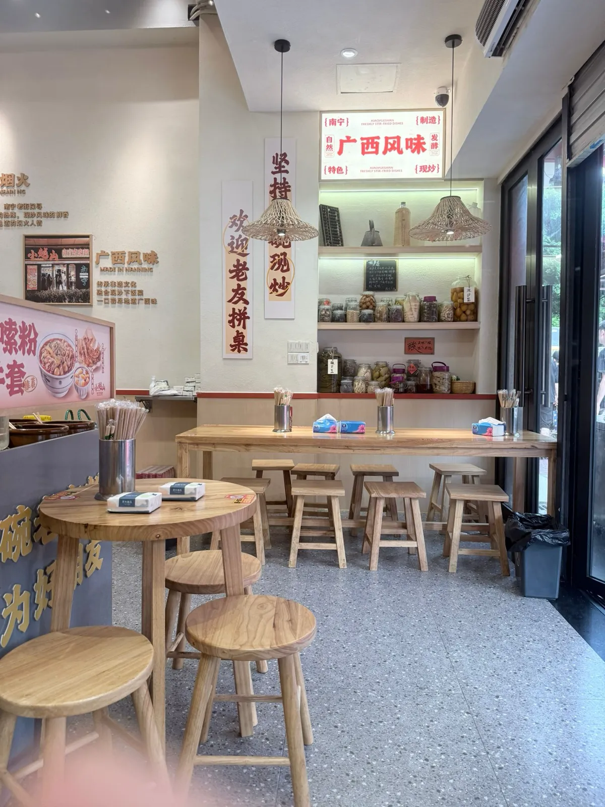 店內堂食區：淺色木長枱同一排矮木凳，枱上放住不銹鋼筷子筒同紙巾；後面牆身內嵌木層架，一排排玻璃罐裝住醃菜同醃筍，上面掛住紅字燈箱寫「廣西風味」，旁邊兩幅直幡寫「堅持碗碗現炒」同「歡迎老友拼桌」；左邊牆掛住一個相框，寫住「廣西風味 MADE IN NANNING」