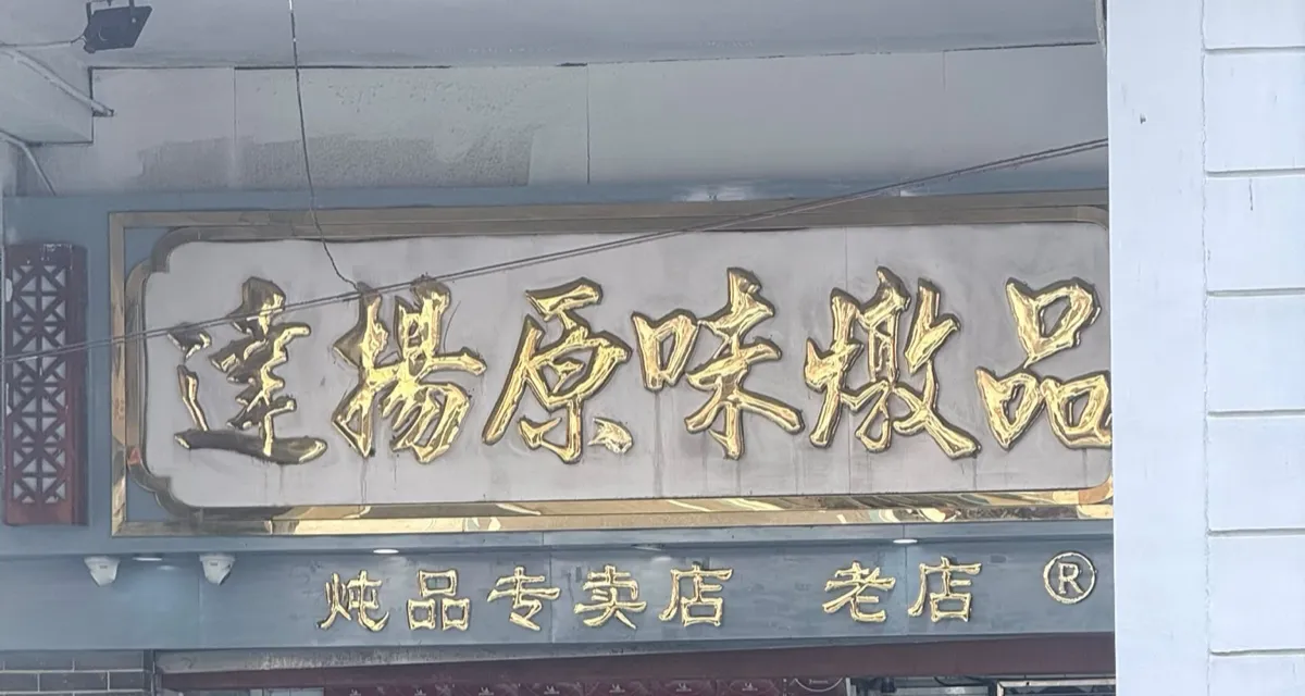 店面招牌：一塊金色立體大字木牌鑲喺淺灰石框入面，寫住「達楊原味燉品」，最右邊個「品」字俾旁邊嘅樓柱遮住一半；招牌下面一條藍灰色橫樑，同樣用金字寫住「炖品专卖店　老店」，後面跟住一個註冊商標圓圈 R，橫樑底掛住兩盞小射燈