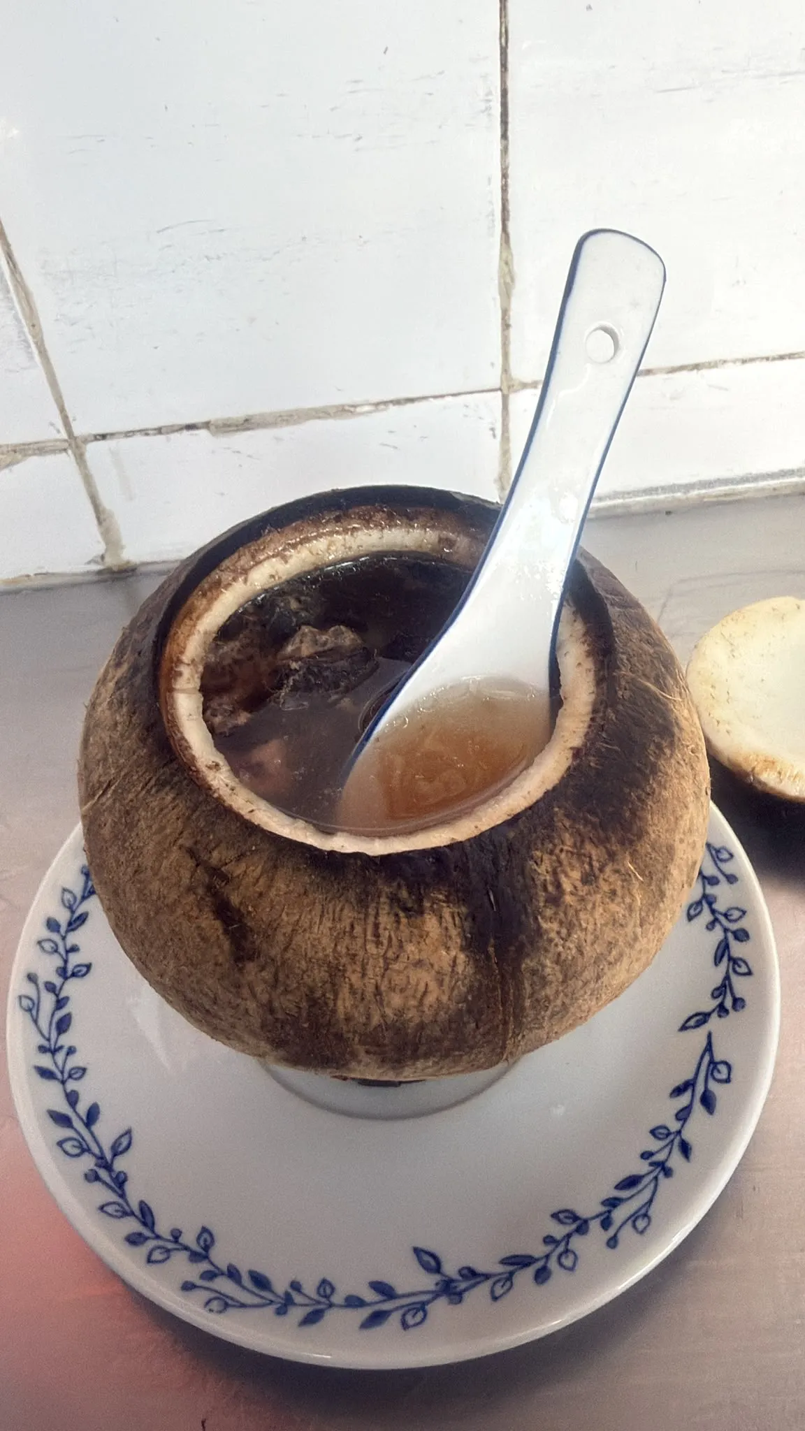 同一個原隻椰子開咗蓋：椰肉內壁白色，入面盛住淺褐色嘅清湯，湯面浮住深色嘅雞肉塊，一隻白底藍邊瓷匙羹插喺湯度；椰子坐喺白底藍色花草邊瓷碟上，右後方放住已經揭起嘅椰蓋，背景係白色方磚牆