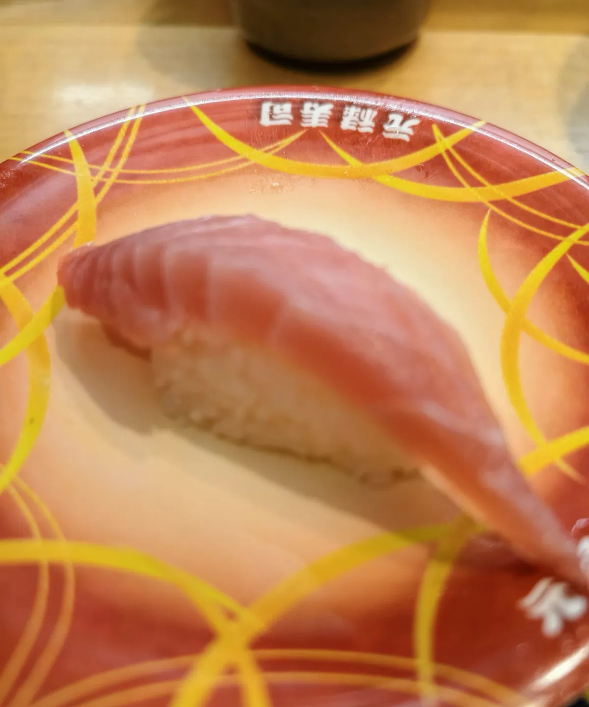 一隻紅色壽司碟，碟面有黃色弧線花紋，碟邊用白字印住「元祿寿司」；碟中間一件握壽司，面頭係一片深粉紅色嘅魚生，見到一條條紋理，下面係白色醋飯；背景係木枱同一隻深色茶杯