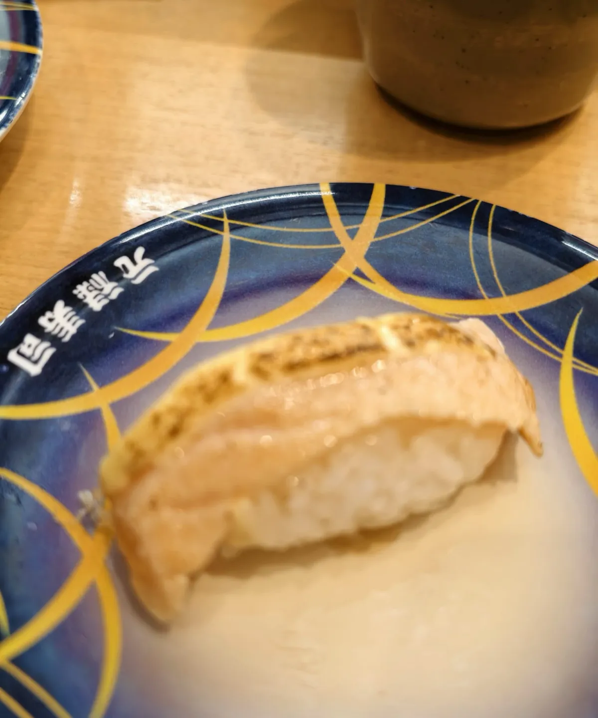 一隻深藍色壽司碟，碟面有黃色弧線花紋，碟邊用白字印住「元祿寿司」；碟上一件握壽司，面頭嗰片淺橙色魚生表面有一行行燒焦咗嘅炙痕，下面係醋飯；背景係木枱同一隻深色茶杯