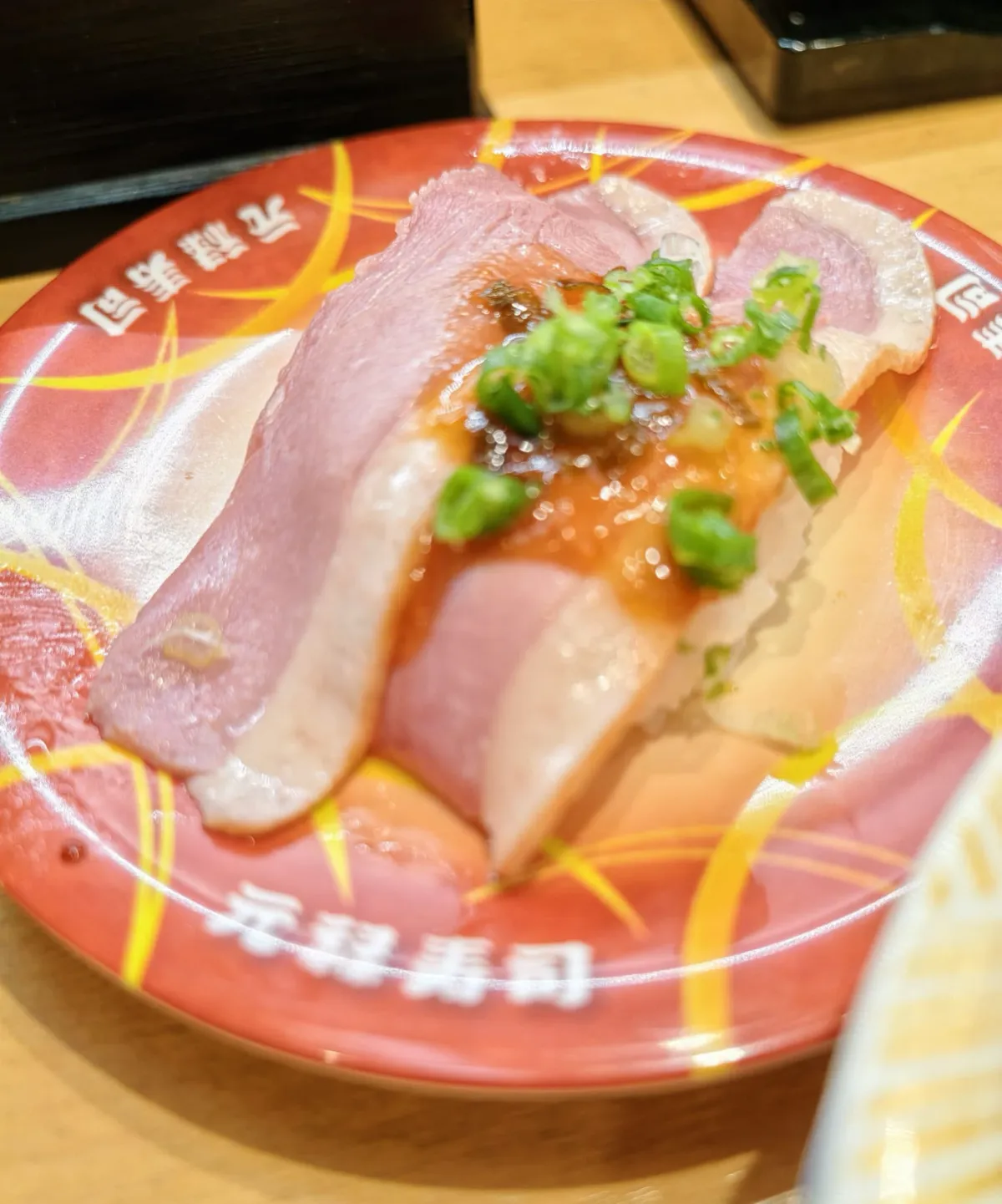 一隻紅色壽司碟，碟邊用白字印住「元祿寿司」；碟上一件握壽司，兩片粉紅色魚生疊喺醋飯上，魚生邊位顏色較淺，面頭淋咗一層褐色醬汁，灑咗一撮青蔥粒；右下角露出另一隻碟嘅邊