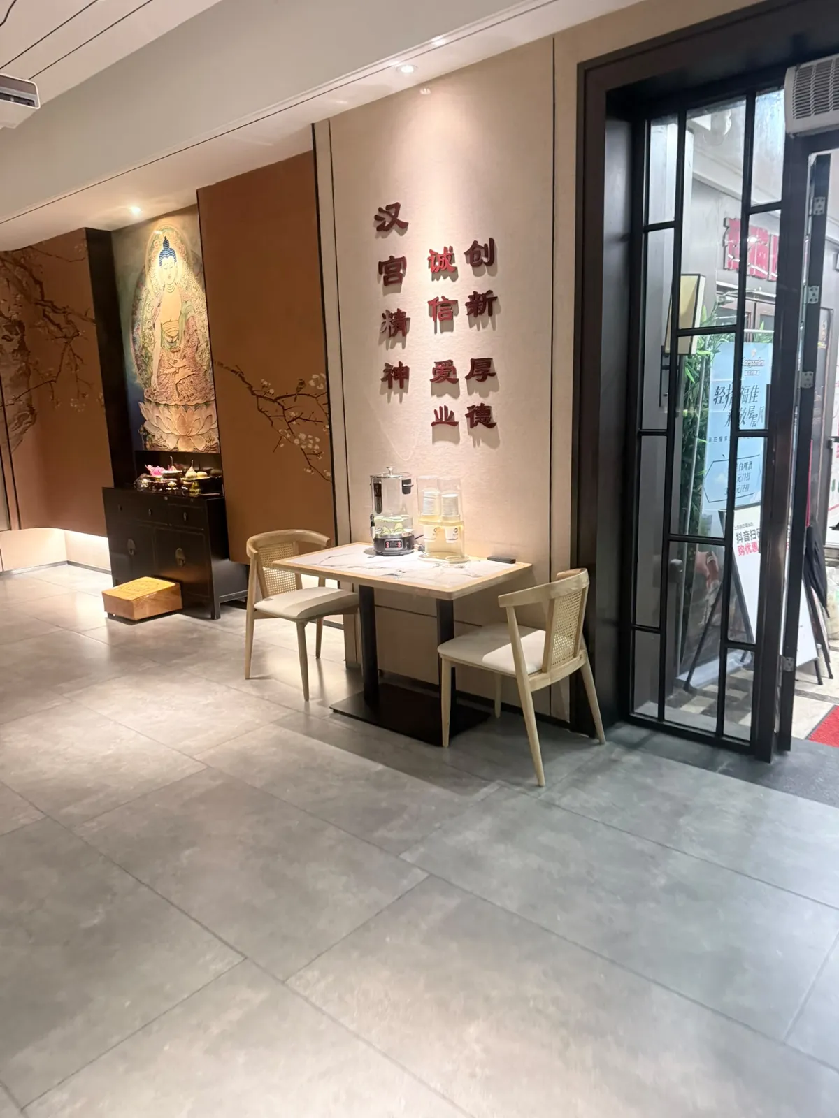 按摩店大堂：米色牆身掛住深色立體中文字，可見「漢宮」同「創新」「厚德」等字樣；左邊牆上有一幅人像工筆掛畫同一張深木供枱，枱上有小擺設；中央一張淺木細枱配兩張藤背椅，枱面放住玻璃水樽同杯；右邊係黑框玻璃門，門外見到街景