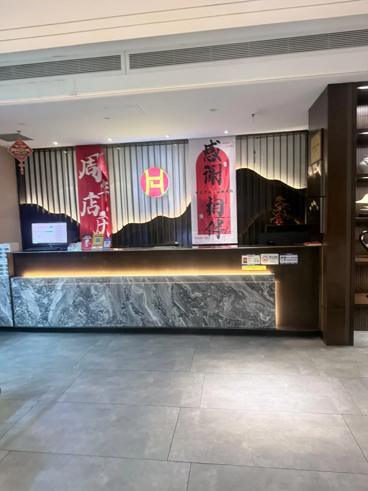 按摩店接待前枱：一列灰白雲石紋長櫃枱，枱面擺住電腦螢幕同幾張小告示牌；背後牆身有直紋木飾同深色山形圖案，掛住兩幅紅色直幅，字樣包括「周年店慶」同「感謝相伴」，中央有一個紅底金字圓形標誌