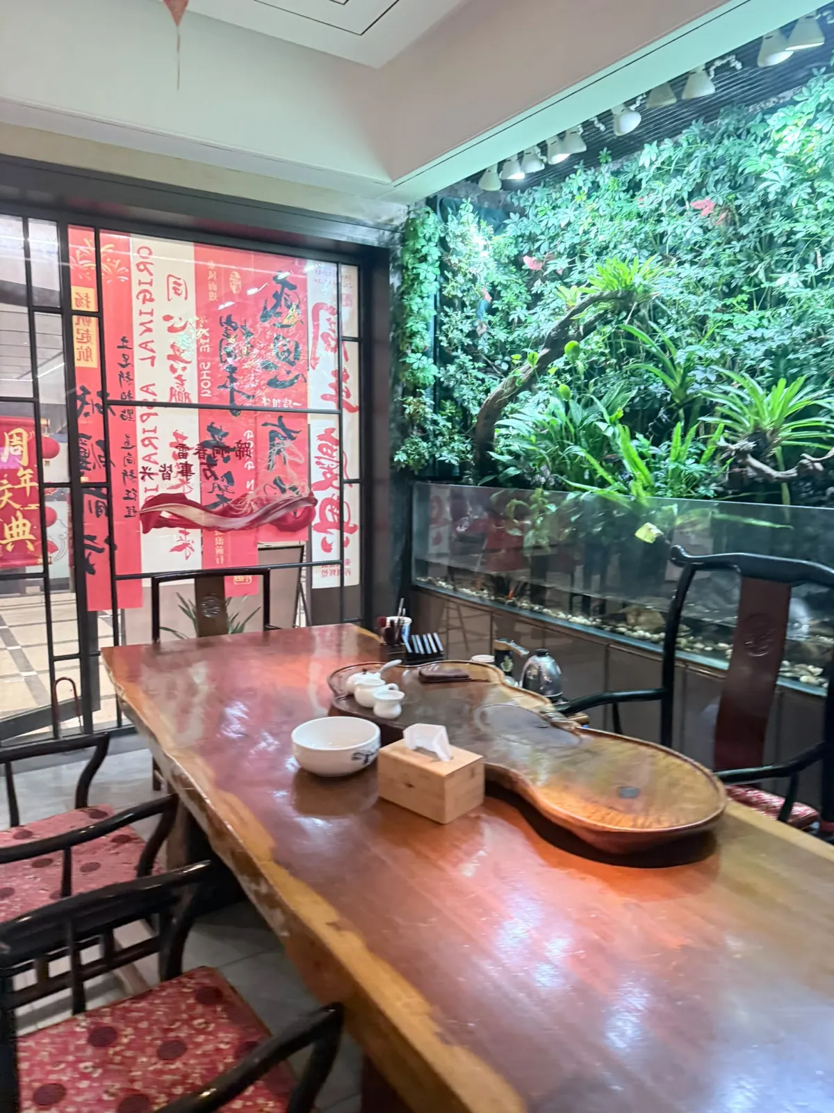 室內休息區：一張厚身原木大板長枱，枱面放住茶盤、茶具、白瓷小碗同一個木紙巾盒，兩旁係深色中式圈椅配紅花座墊；右邊整幅牆係綠色植物牆，下方有一個長形玻璃缸；左邊玻璃窗貼滿紅色書法海報，可見「週年慶典」字樣