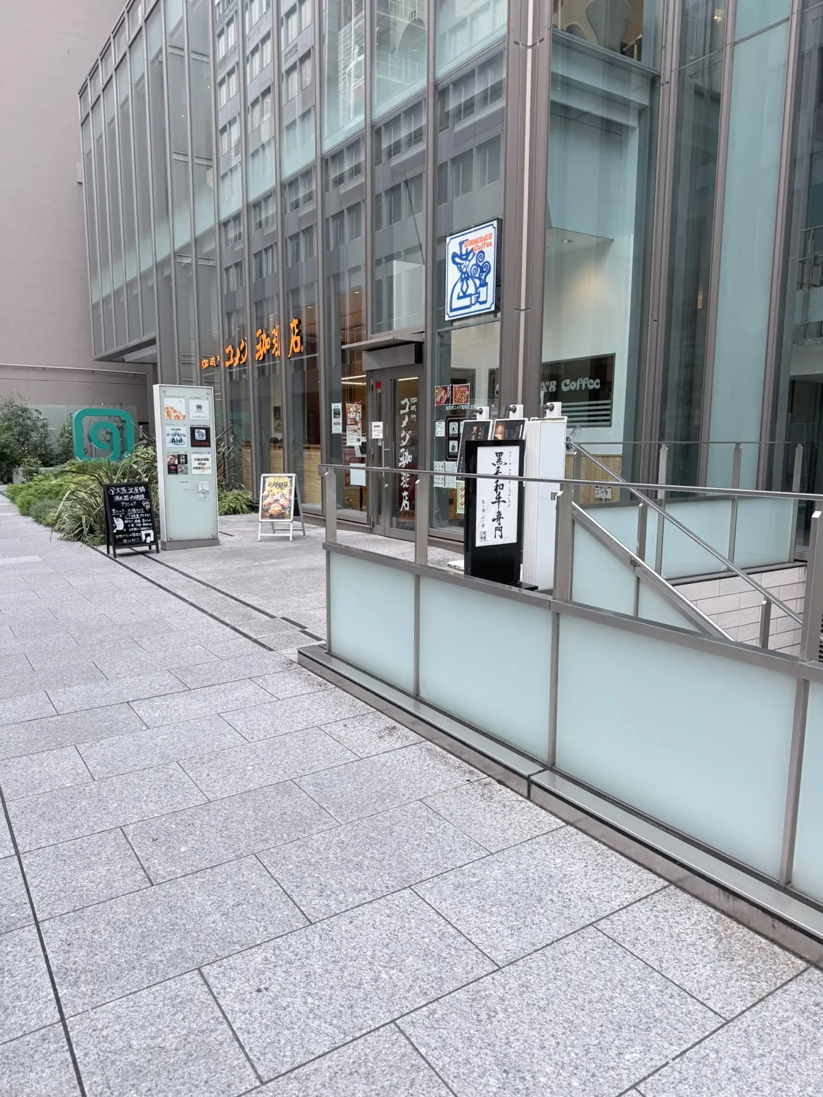 一座玻璃幕牆大廈嘅地下：玻璃上面有一行橙色大字「コメダ珈琲店」，門口上方掛住一塊藍白色方形招牌，畫住一個戴高帽嘅卡通人物；玻璃門旁邊有一塊直排店名牌，門前擺住一塊食物圖片嘅企牌；左邊有綠化帶同大廈嘅租戶指示牌，右邊有欄杆同另一塊黑色直立招牌；前景係一大片灰色石磚行人路