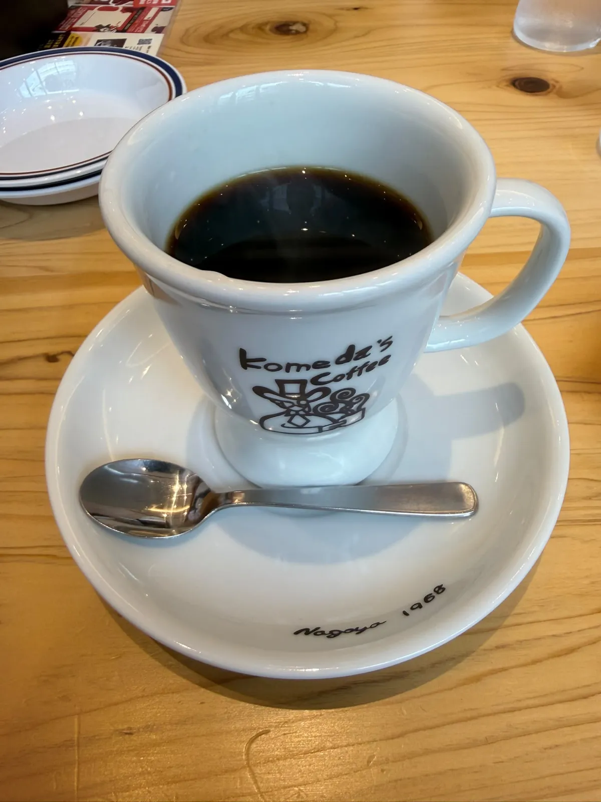 木枱上一隻白色高腳咖啡杯，杯身用啡色印住「Komeda's Coffee」同一個戴高帽嘅卡通人物，杯入面係一杯黑咖啡；杯放喺一隻白色深碟上，碟上擺住一支金屬茶匙；左上角見到一疊藍紅邊白碟同一張紙餐墊嘅一角，右上角有一杯冰水
