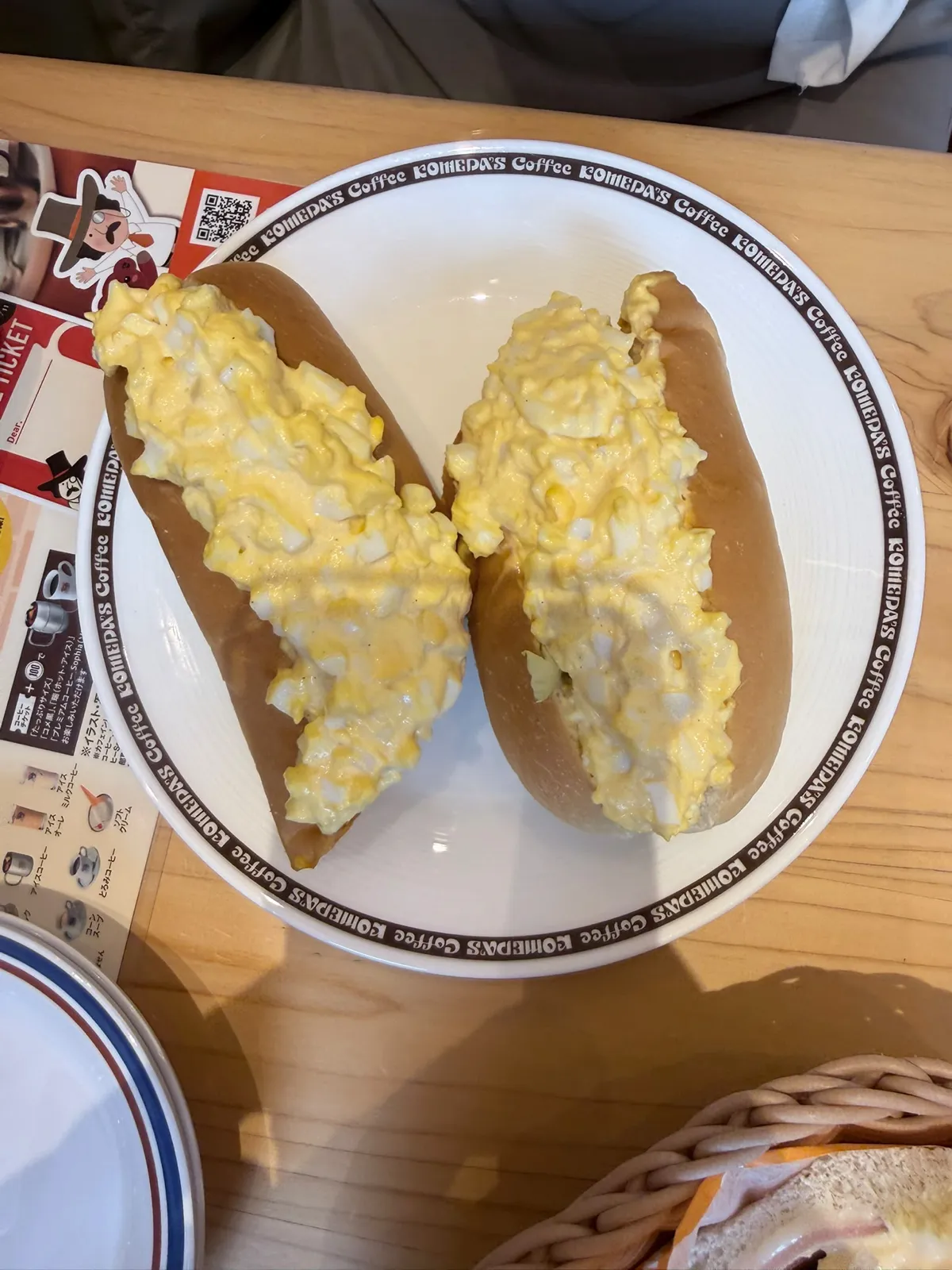 由上向下影嘅一隻白碟，碟邊一圈啡色字印住「KOMEDA'S Coffee」；碟上兩條切開嘅軟身長麵包，中間夾滿淡黃色嘅雞蛋沙律，滿到瀉出麵包面；左邊壓住一張印有卡通插畫同 QR code 嘅紙餐墊，最頂見到同枱一位人士嘅灰色衫，冇臉入鏡；左下角同右下角分別露出另一隻碟同一個籐籃嘅邊