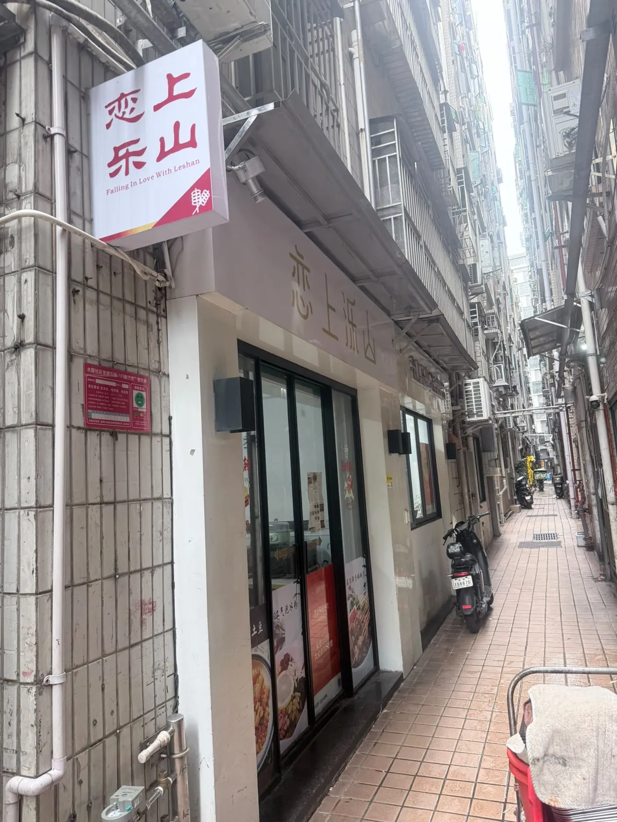 一條被兩邊高樓夾住嘅窄巷：左邊係鋪米色瓷磚嘅唐樓外牆，牆上伸出一個白底紅字嘅方形吊牌，直排寫住「戀上樂山」，下面一行細英文 Falling In Love With Leshan，右下角有個紅色串燒圖案；吊牌後面係店舖嘅淺灰色雨篷，雨篷側面用同色暗字再寫一次店名；店面係黑框玻璃趟門，門邊貼住幾張食物圖片；巷嘅右邊泊住一部電單車，地面鋪淺啡色地磚
