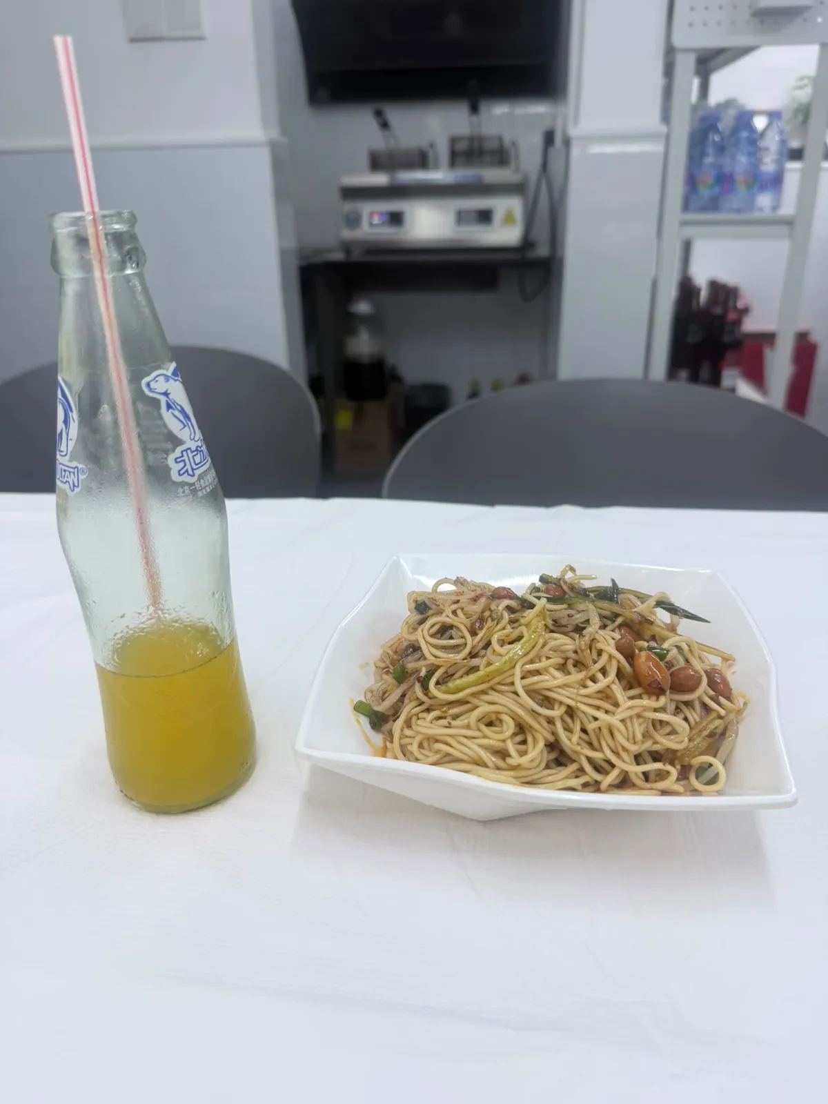 白枱布上面擺住一支玻璃樽汽水同一碟麵：樽身結滿水珠，樽貼上面係一隻藍色北極熊同「北冰洋」三個字，入面淨返大約三分一橙黃色飲品，插住一支紅白間條飲管；右邊白色方碟盛住一堆淺黃色麵條，面頭見到芽菜、青瓜條、青蔥段同幾粒炸花生，麵身掛住褐色醬汁；背景係店內開放式廚房，見到不鏽鋼工作枱、電磁爐同一排樽裝水