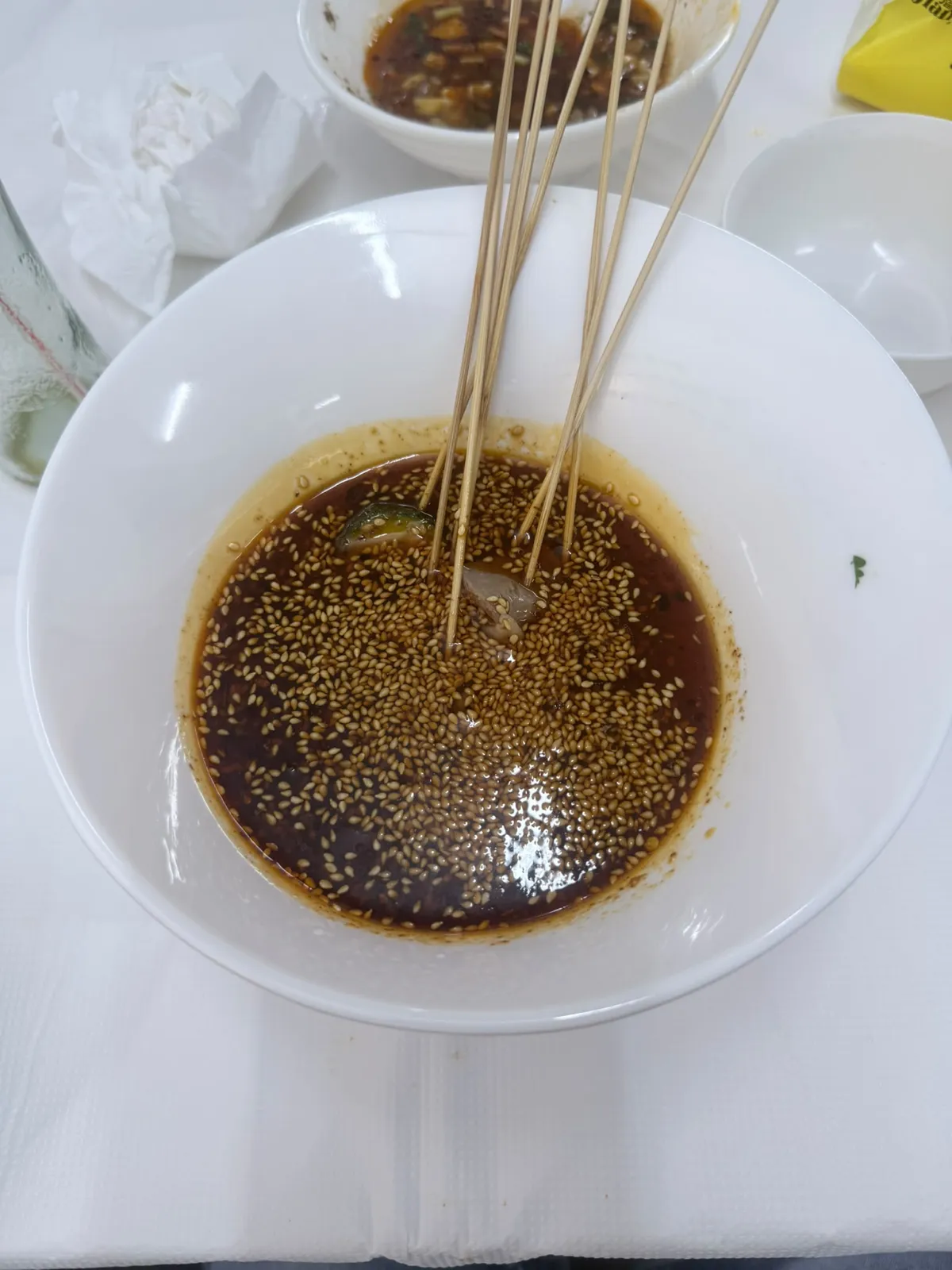 一隻白色深碗盛住深褐色嘅紅油蘸汁，汁面浮住厚厚一層白芝麻，見到一小塊青色辣椒同一塊淺色肉；一束用完嘅幼竹籤斜插喺碗入面，向右上方伸出碗口；碗擺喺白色枱布上，後面另一隻白碗入面剩返深色湯汁同蔥花，右上角有一疊乾淨嘅白碗