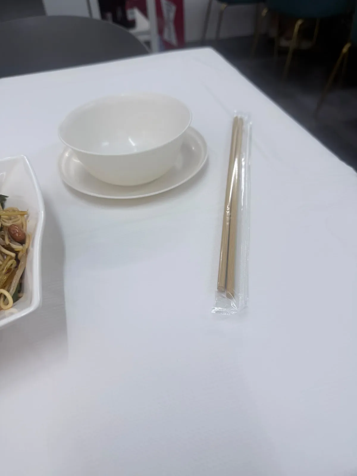 白枱布上面擺住一隻白色瓷碗，碗坐喺一隻白色小碟上；右邊平放住一對未拆封嘅竹筷子，連住透明膠袋包裝；左下角見到另一隻白色方碟嘅一角，入面有麵條同炸花生；背景見到店內嘅椅腳同一個紅色紙箱