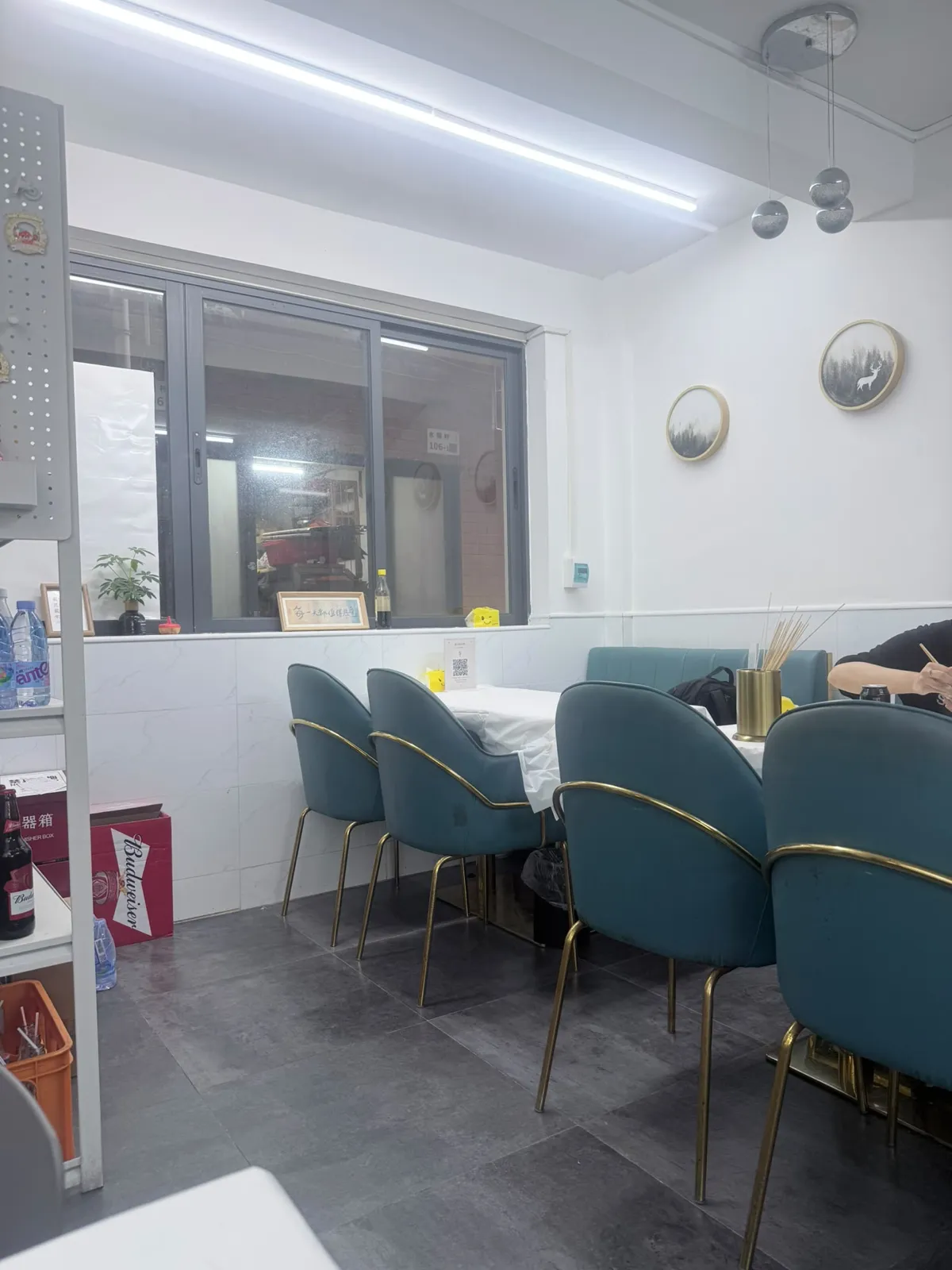 店內用餐區：白色天花吊住一支長條光管，牆身下半鋪白色雲石紋磚；灰色鋁窗外面望到巷仔，窗框上貼住一塊寫住「水圍村 106-1」嘅門牌；右邊白牆掛住兩幅圓形金框風景畫；窗下一行四張墨綠色絨面椅，配金色幼椅腳，圍住兩張鋪咗白布嘅枱，枱上放住一個金色圓筒，入面插住一紮竹籤；畫面最右有一位食客，得手臂同上身入鏡；左邊層架擺住樽裝水同汽水，地下放住一個紅色紙箱