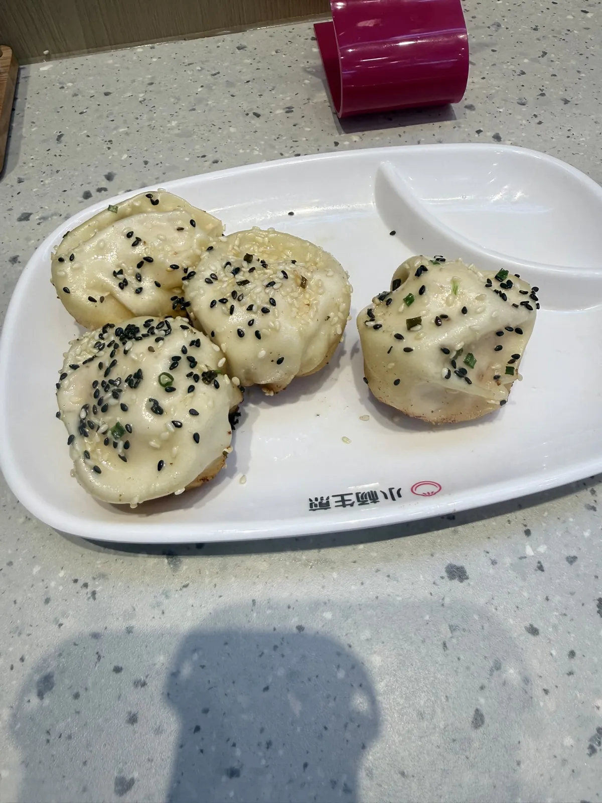 麻石紋枱面上一隻白色橢圓分格碟，左格排住四隻生煎包，包面呈奶白色、頂部撒滿黑白芝麻同青蔥碎，底部露出金黃煎面；右格空住留俾點醬，碟身印住「小杨生煎」字樣