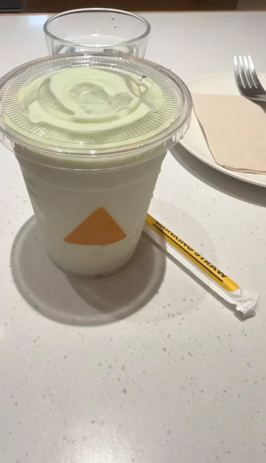 一杯淺綠色飲品，透明蓋，杯身有橙色三角標記，杯外有水珠；旁邊一條未拆嘅黃色飲管