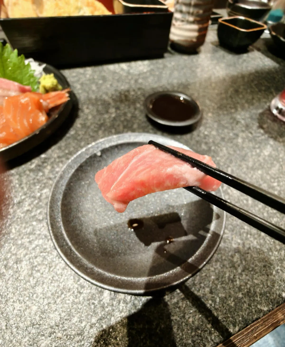 黑色筷子夾住一片粉紅色、有白色油脂紋嘅魚生，下面係一隻淺淺醬油嘅黑碟；左邊碟邊見到三文魚同蝦